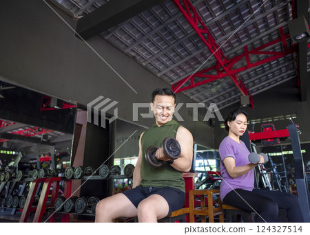 Indonesian southeast asian man and chinese east asian woman working out lifting a dumbbell at the gym. Fitness exercise and healthy lifestyle concept Indonesian southeast asian man and chinese east asian woman working out lifting a dumbbell at the gym. Fitness exercise and healthy lifestyle concept 124327514