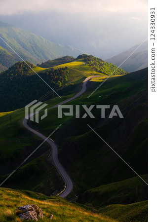 View of mountain road in fields at sunset in summer. Beautiful landscape with winding road among green meadows, golden sunlight in summer. Travel View of mountain road in fields at sunset in summer. Beautiful landscape with winding road among green meadows, golden sunlight in summer. Travel 124330931