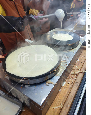 A person pouring a ladle of batter into a round pan for frying pancakes. A cook making pancakes from fried dough. Frying pancakes outdoors. Cooking street food at festival. Cooking meals on the street A person pouring a ladle of batter into a round pan for frying pancakes. A cook making pancakes from fried dough. Frying pancakes outdoors. Cooking street food at festival. Cooking meals on the street 124331644