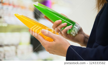 A female customer compares two tubes of shower gel or facial cleanser, reading label with ingredients and choosing between them for a conscious decision in the cosmetic department. Skincare products. 124333582