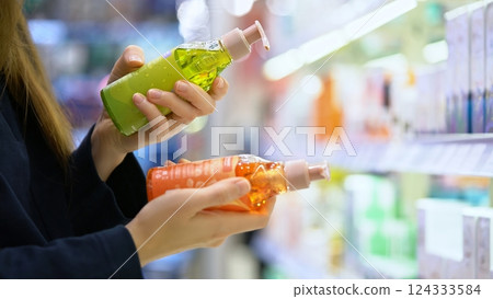 A female customer compares two shampoo different bottles for hair care, reading ingredient labels and examining composition for self care. Conscious choice. Close-up of hands in a cosmetic department. A female customer compares two shampoo different bottles for hair care, reading ingredient labels and examining composition for self care. Conscious choice. Close-up of hands in a cosmetic department. 124333584