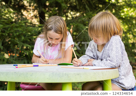 Two children sketching with colorful pens at a table in a green outdoor setting Two children sketching with colorful pens at a table in a green outdoor setting 124334654