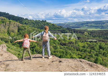 Two young girls with backpacks standing on rocky cliff with arms outstretched overlooking forest valley Two young girls with backpacks standing on rocky cliff with arms outstretched overlooking forest valley 124334655