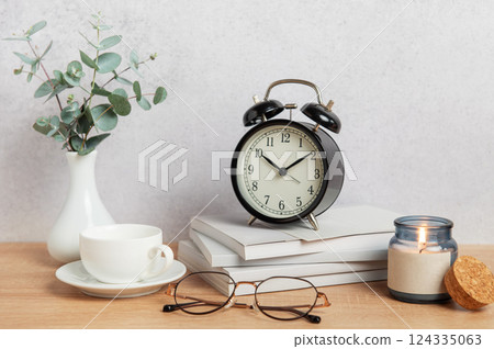 Alarm clock, books, coffee cup, eyeglasses and candle creating cozy atmosphere on wooden desk 124335063