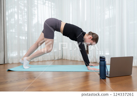 Young woman practicing yoga indoors with online guidance using laptop. Woman doing stretching exercises for flexibility on yoga mat. Concept of home fitness, flexibility, and wellness. 124337261