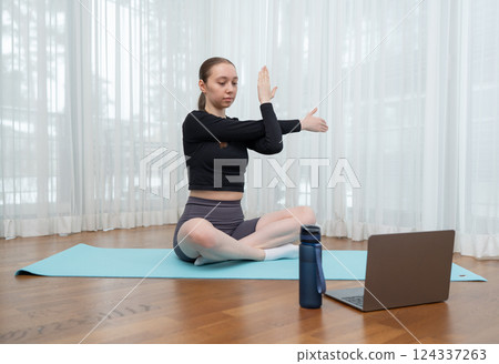 Young woman stretching on a yoga mat while watching an online workout on laptop, with copy space. Concept of fitness, flexibility, and virtual training Young woman stretching on a yoga mat while watching an online workout on laptop, with copy space. Concept of fitness, flexibility, and virtual training 124337263
