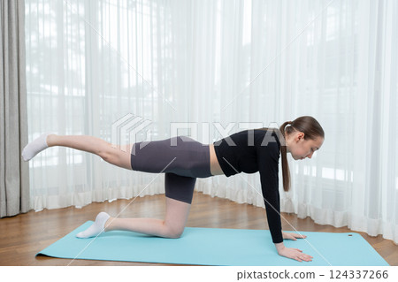 Young woman practicing yoga at home on a blue yoga mat. Balance and stretching exercises at home. Concept of balance, strength, and mindfulness in a comfortable indoor environment. 124337266