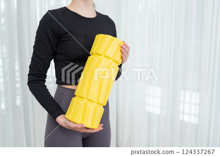 Fit woman holding a yellow massage foam roller for muscle recovery wearing workout clothes. Concept of fitness equipment, body care, and post-workout therapy. 124337267