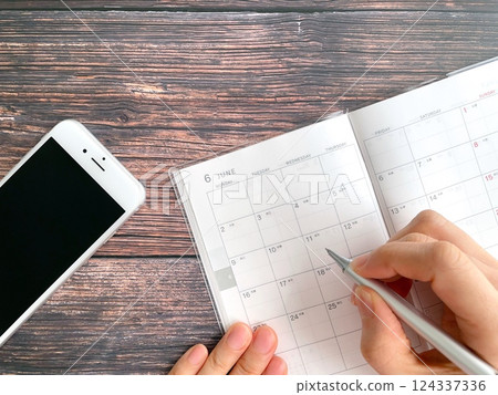 一位女士在寫 6 月份日程表的手和桌上的智慧型手機 124337336