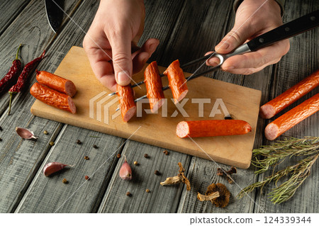 A person carefully skewers a smoked sausage on a wooden cutting board. Garlic, dried herbs and spices are arranged around the board, adding to the rustic culinary atmosphere A person carefully skewers a smoked sausage on a wooden cutting board. Garlic, dried herbs and spices are arranged around the board, adding to the rustic culinary atmosphere 124339344