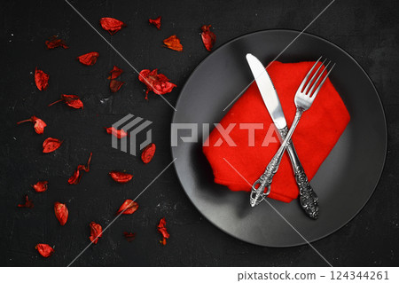 Fork, knife with red napkin, roses and black plate. On dark table background. Fork, knife with red napkin, roses and black plate. On dark table background. 124344261