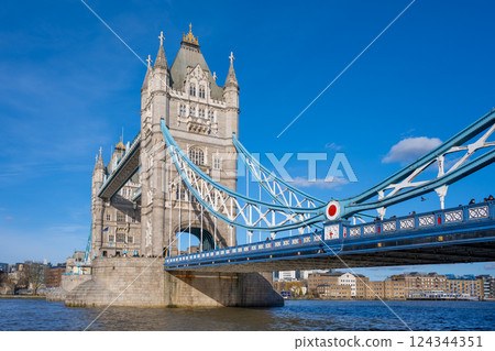 Tower Bridge stands majestically over the River Thames in London, showcasing its intricate architecture and vibrant blue cables against a bright sky, with people walking across. 124344351