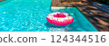Pink donut-shaped inflatable float in turquoise swimming pool with wooden deck in sunny backyard 124344516
