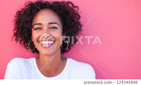 Portrait of a joyful young woman with curly hair and hoop earrings laughing in front of a vibrant pink wall, wearing a white top and radiating positive energy. 124344806