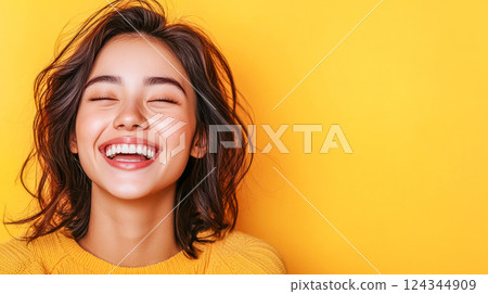 Portrait of a cheerful young woman with long dark hair and a wide smile, wearing a yellow sweater against a vibrant yellow background with copy space on the right. 124344909