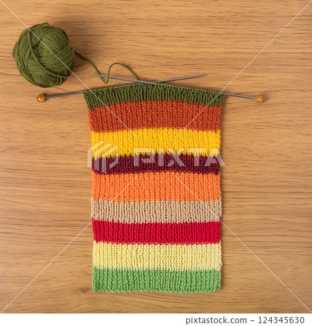 A striped knitted fabric with knitting needles and a green knitting ball on a brown wooden table. Knitting striped bright scarf made of red, yellow, green, orange and purple threads. Knitted warm 124345630