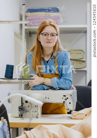 Woman stands in front of a sewing machine, wearing a yellow apron. Woman stands in front of a sewing machine, wearing a yellow apron. 124345810