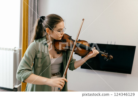 Young woman playing violin at home by sheet music, focused on learning music. Violin student rehearsing classical music in a professional study session Young woman playing violin at home by sheet music, focused on learning music. Violin student rehearsing classical music in a professional study session 124346129