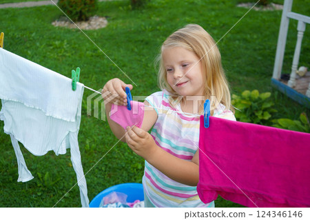 A happy six-year-old stands beside a swaying clothesline, securing each piece with care while enjoying the warm breeze. 124346146