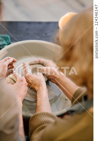 Elderly Caucasian woman practices shaping clay on a pottery wheel at a master class. A young instructor guides her hands, teaching sculpting techniques at a creative workshop 124346147