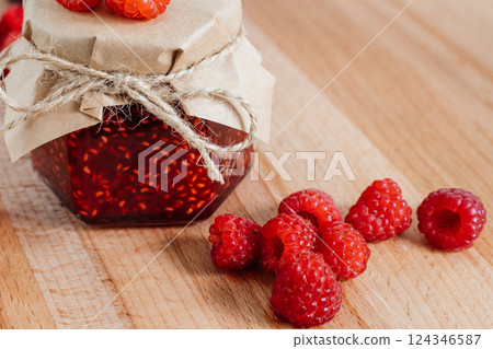 Raspberry jam in a jar on a wooden board, the lid is decorated with paper and tied with twine. Raspberries are lying nearby. Banner, menu, recipe place for text 124346587