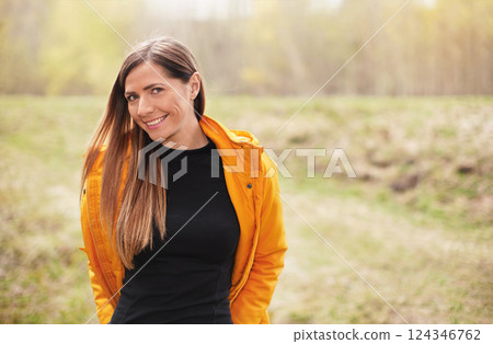 Portrait of young woman in yellow jacket, smiling, long hair down, blurred trees background Portrait of young woman in yellow jacket, smiling, long hair down, blurred trees background 124346762
