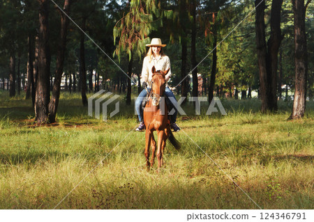 Young woman in shirt and straw hat, riding brown horse in the park, blurred background with houses and trees 124346791