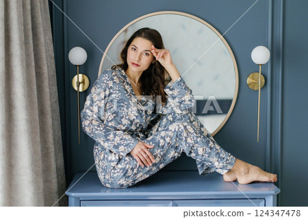 Woman wearing pajamas sitting on furniture in bedroom with round mirror and wall lamps 124347478