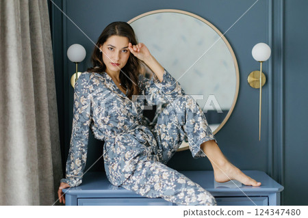 Woman wearing pajamas sitting on furniture in bedroom with round mirror and wall lamps 124347480