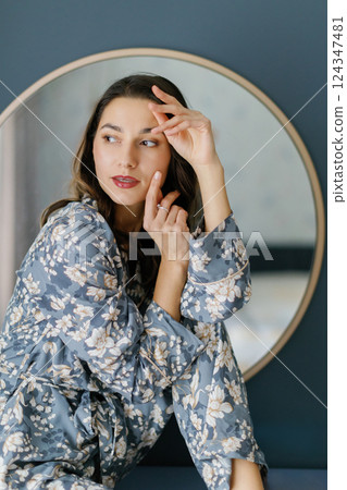 Woman wearing pajamas sitting on furniture in bedroom with round mirror and wall lamps Woman wearing pajamas sitting on furniture in bedroom with round mirror and wall lamps 124347481