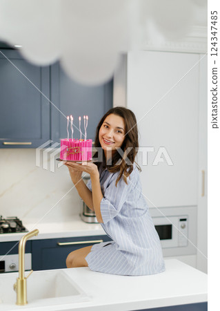 Woman celebrating birthday holding pink cake with candles in modern kitchen Woman celebrating birthday holding pink cake with candles in modern kitchen 124347485