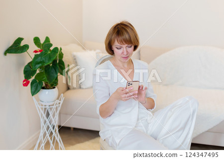Adult beautiful woman relax siting on chair at home and using cellphone texting chatting with friend. Happy young woman on chair holding smartphone in her hands, shoping online or check social media. 124347495