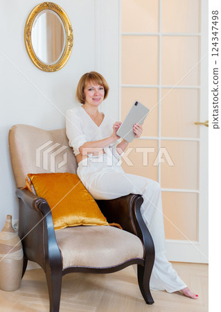 Smiling mature woman holding tablet, relaxing on a armchair at home in the living room. 124347498