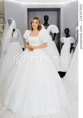 Pretty young woman is choosing a wedding dress in the shop. The bride-to-be is wearing a wedding dress for fitting Pretty young woman is choosing a wedding dress in the shop. The bride-to-be is wearing a wedding dress for fitting 124347602