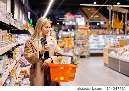 Woman choosing canned food, holding shopping basket in supermarket Woman choosing canned food, holding shopping basket in supermarket 124347881