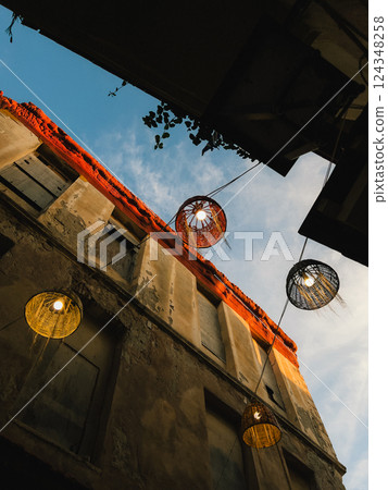 Wicker lamps illuminating old building facade. Low angle view of traditional lamps hanging between buildings in narrow street of mediterranean town, creating warm and inviting atmosphere at sunset Wicker lamps illuminating old building facade. Low angle view of traditional lamps hanging between buildings in narrow street of mediterranean town, creating warm and inviting atmosphere at sunset 124348258