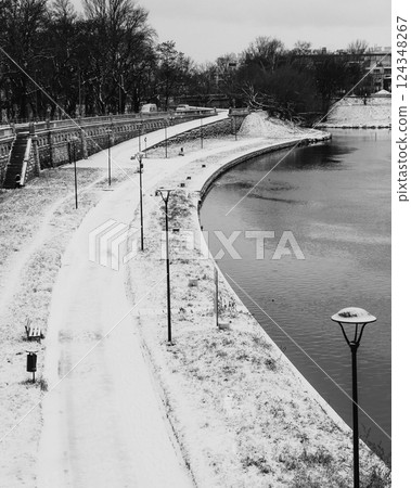 Snow covered promenade lining the river in winter, featuring street lamps, benches, and trash cans, captured in striking black and white image that evokes tranquility and solitude. promenade in winter Snow covered promenade lining the river in winter, featuring street lamps, benches, and trash cans, captured in striking black and white image that evokes tranquility and solitude. promenade in winter 124348267