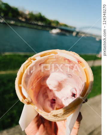 Hand holding melting strawberry ice cream. Tourist savoring a melting strawberry ice cream cone by the river, enjoying the warmth of a sunny summer day while surrounded by picturesque urban landscape 124348270