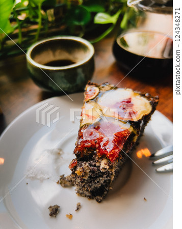 Poppy seed cake with fig slices and berry or fruit glaze on a white plate, partially eaten with a fork, accompanied by a cup of coffee or tea and a coffee pot on a wooden table. First-person view 124348271