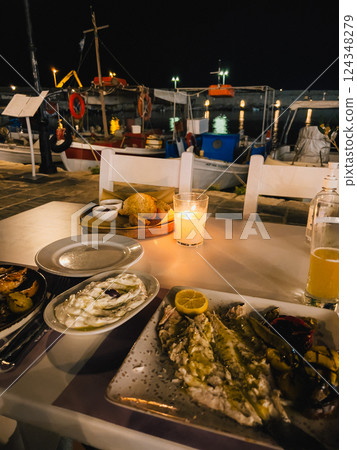 First-person view: Plates of fresh fish, grilled vegetables are served on table at outdoor restaurant by harbor at night, illuminated by candle, creating cozy and inviting atmosphere. greek dinner 124348279