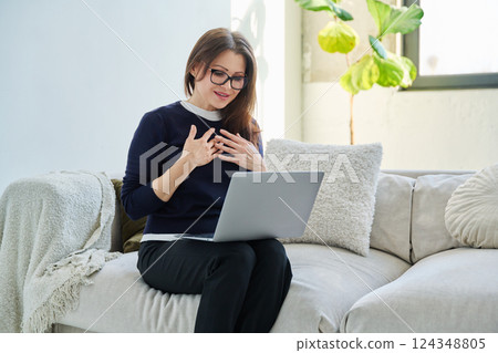 Middle aged woman sitting at home on sofa using laptop for video call, chat 124348805