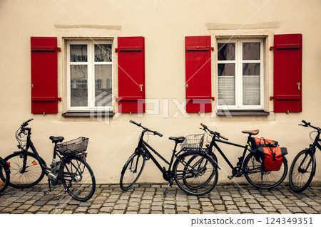 A picturesque European facade with two windows featuring red wooden shutters. Parked bicycles along 124349351