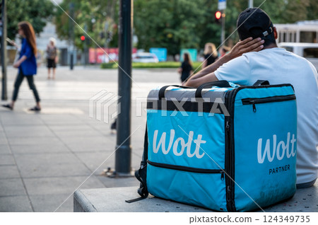 Budapest, Hungary, August 30, 2022. A worker at Wolt, a food delivery company. He is taking a break sitting with his light blue delivery bag and logo next to him. Gig economy. Budapest, Hungary, August 30, 2022. A worker at Wolt, a food delivery company. He is taking a break sitting with his light blue delivery bag and logo next to him. Gig economy. 124349735