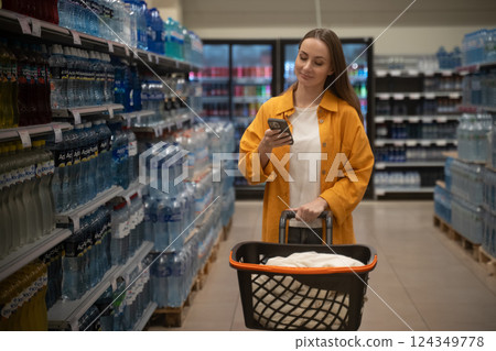 Woman shopping in supermarket aisle while checking her phone for grocery list Woman shopping in supermarket aisle while checking her phone for grocery list 124349778