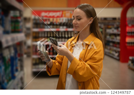 Woman uses smartphone to scan household cleaning products aisle at grocery store during daylight, looking for deals 124349787