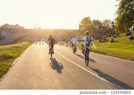 Scenic view youth people ride bicycles sunny Dresden Elbe riverside path against warm sunset glow active leisure. Rider perform wheelie beside historic Augustus bridge crowd relaxes grass lawn meadow 124349905
