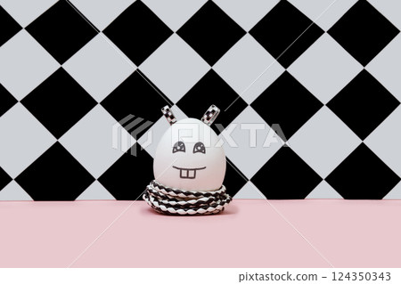 A creative cheerful Easter white egg in the form of a rabbit in black and white style with a painted muzzle, eyes and bunny smile with two teeth and ears. Stylized funny character on chess background. A creative cheerful Easter white egg in the form of a rabbit in black and white style with a painted muzzle, eyes and bunny smile with two teeth and ears. Stylized funny character on chess background. 124350343