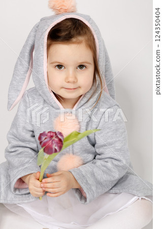 Curious Toddler in Bunny Costume Looking at a Single Burgundy Tulip on White Background 124350404