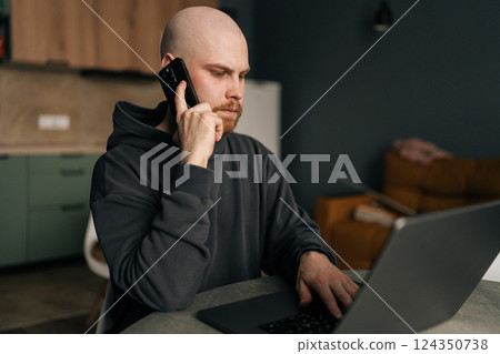 Serious freelancer male working from remote home office, talking on mobile phone and using laptop computer, managing business and multitasking in home environment. Concept of remote job, technology. 124350738