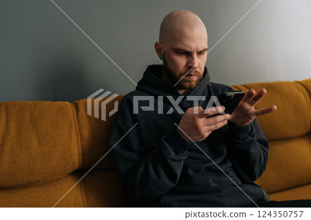 Close-up of serious young bald man with beard and hoodie using smartphone while sitting on comfortable sofa, browsing internet, social media or mobile application. Concept of modern technology. Close-up of serious young bald man with beard and hoodie using smartphone while sitting on comfortable sofa, browsing internet, social media or mobile application. Concept of modern technology. 124350757